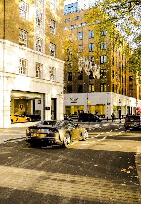 A city street scene in Grosvenor Square, Mayfair, featuring a row of multi-storey buildings with large windows, some with retail stores on the ground floor. The buildings are constructed with a combination of light-colored stone and darker brickwork. In the foreground, a sleek black sports car is parked near the curb, while additional cars, including a yellow sports car visible through the glass storefront, are parked along the street. The pavement shows clearly marked pedestrian crossings, and a few people are walking on the sidewalk. Mature trees with green and yellow foliage line the street, casting dappled sunlight on the scene. Brightly lit signage and reflections on glass windows highlight the commercial environment. This urban setting emphasizes cleanliness and maintenance of the premises, aligning with the professional cleaning services offered by Carpet Cleaners Mayfair.