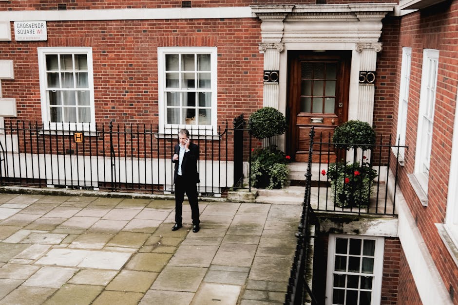 A city street scene in Grosvenor Square, Mayfair, featuring a row of multi-storey buildings with large windows, some with retail stores on the ground floor. The buildings are constructed with a combination of light-colored stone and darker brickwork. In the foreground, a sleek black sports car is parked near the curb, while additional cars, including a yellow sports car visible through the glass storefront, are parked along the street. The pavement shows clearly marked pedestrian crossings, and a few people are walking on the sidewalk. Mature trees with green and yellow foliage line the street, casting dappled sunlight on the scene. Brightly lit signage and reflections on glass windows highlight the commercial environment. This urban setting emphasizes cleanliness and maintenance of the premises, aligning with the professional cleaning services offered by Carpet Cleaners Mayfair.