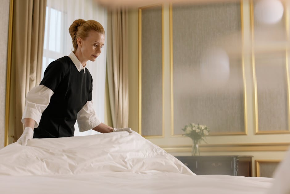 A professional cleaner with neatly styled reddish-brown hair, dressed in a black uniform layered over a white shirt, is carefully making a bed with a white duvet cover in a luxurious hotel room. The room features elegant gold-accented wall panels, soft natural lighting from large windows with beige curtains, and a wooden side table with a vase of white flowers, creating a tidy and pristine environment. Visible cleaning tools are not shown, but the overall scene emphasizes meticulous surface cleaning, deep cleaning, and hygiene practices associated with domestic or hospitality cleaning services, as provided by Carpet Cleaners Mayfair.