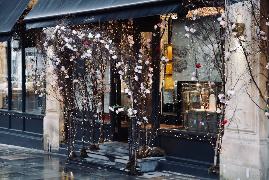 The exterior of a boutique building on Bond Street is decorated with twisted, leafless branches intertwined with string lights and scattered faux blossoms, predominantly in pink and white hues. The entrance features small stone steps leading up to glass doors, framed by a black-painted metal structure supporting the decorative branches and lighting. The shop's large display windows offer a glimpse of the interior, which appears clean and well-maintained. The pavement outside is damp, indicating recent cleaning or rain, with reflections of the string lights visible on the wet surface. This scene exemplifies meticulous surface cleaning and maintenance, with attention to aesthetic presentation typical of luxury retail environments, and reflects the high standards of hygiene and presentation upheld by Carpet Cleaners Mayfair in maintaining properties in this prestigious area.