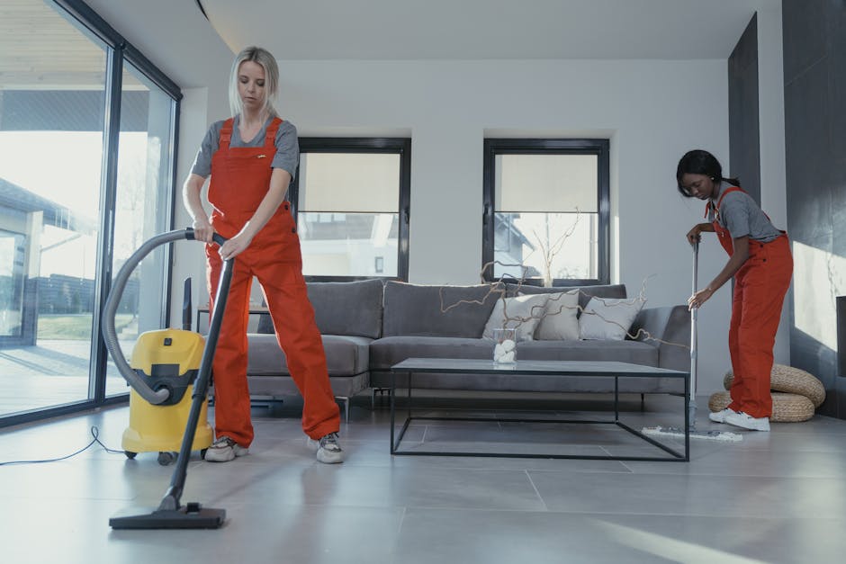 Two professional cleaners from Carpet Cleaners Mayfair, dressed in gray shirts and bright orange overalls, are performing surface cleaning in a modern living room. The cleaner on the left is using a y