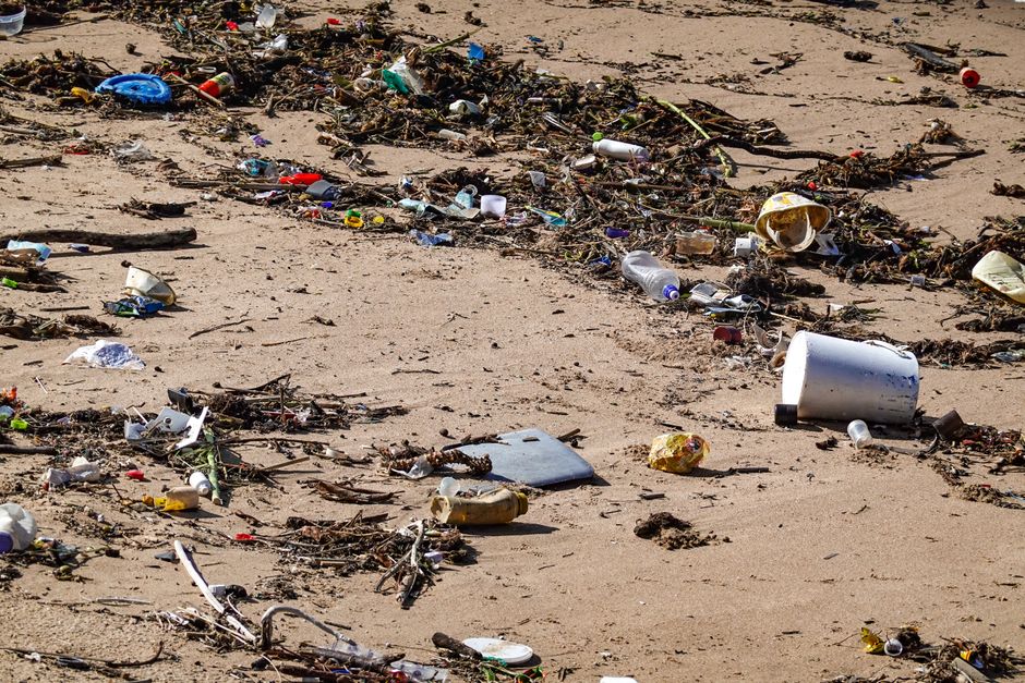 A sandy beach strewn with various types of litter, including plastic bottles, cups, and containers, along with pieces of driftwood and seaweed. The debris is scattered across the light-colored sand, creating a cluttered and unclean appearance. The lighting indicates a bright, sunny day, highlighting the contrast between the natural environment and the man-made waste. This image emphasizes the importance of proper waste disposal and environmental maintenance, concepts relevant to hazardous waste management and cleaning services provided by companies like Carpet Cleaners Mayfair, especially concerning legal obligations around waste disposal.