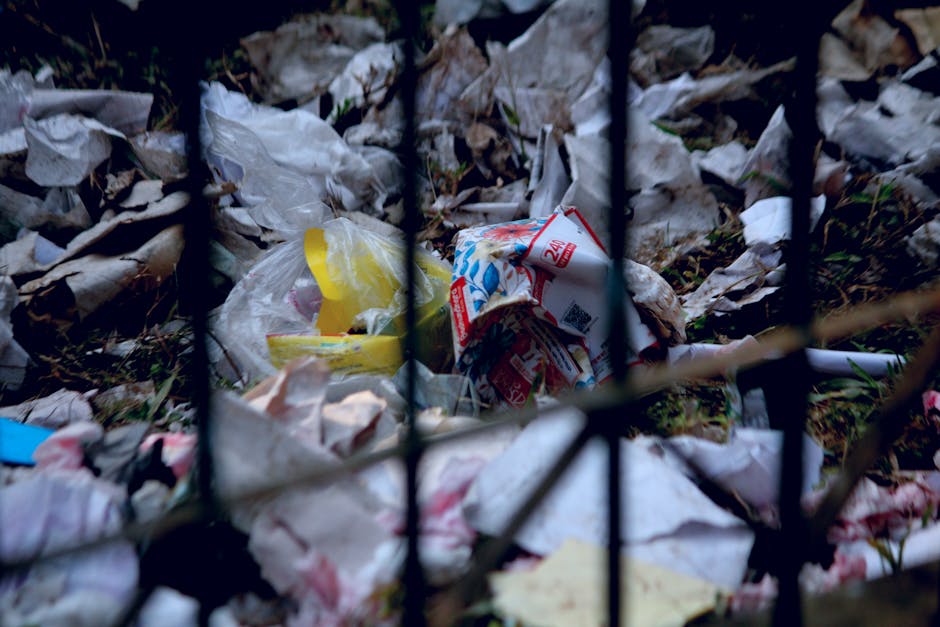 A sandy beach strewn with various types of litter, including plastic bottles, cups, and containers, along with pieces of driftwood and seaweed. The debris is scattered across the light-colored sand, creating a cluttered and unclean appearance. The lighting indicates a bright, sunny day, highlighting the contrast between the natural environment and the man-made waste. This image emphasizes the importance of proper waste disposal and environmental maintenance, concepts relevant to hazardous waste management and cleaning services provided by companies like Carpet Cleaners Mayfair, especially concerning legal obligations around waste disposal.