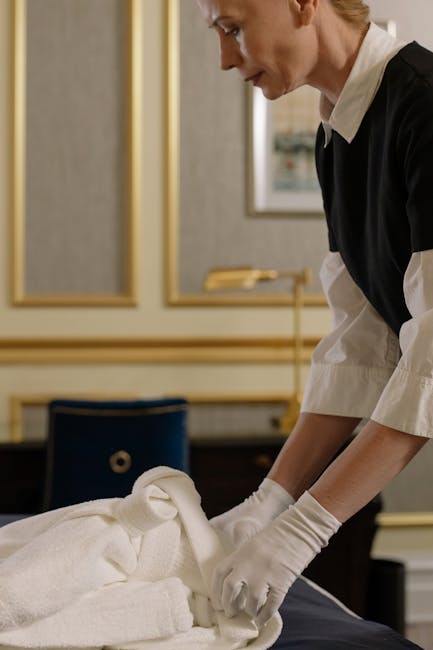 A professional cleaner, dressed in a black and white uniform with white gloves, carefully folds and arranges white towels on a hotel or residential bed in a well-lit room. The room features elegant decor, with framed artwork on the wall and a dark wooden desk or side table partially visible in the background. The surface of the bed appears clean and neatly made, indicating thorough deep cleaning and surface sanitation. Visible tools include the towels being arranged, while the lighting highlights the spotless, fresh appearance of the linens and surrounding area. Carpet Cleaners Mayfair specializes in domestic cleaning and surface sanitation, ensuring hygiene and cleanliness for move-out or regular property maintenance.