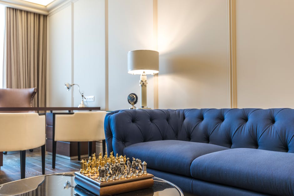 A luxurious Mayfair flat living room featuring a deep blue tufted sofa with a clean, polished surface, positioned next to a beige armchair. The room has light-colored walls with elegant molding and warm wooden flooring. A modern white lamp with a metallic base stands beside the sofa, casting soft light onto the space. In front of the sofa, a glass coffee table holds a chessboard with chess pieces. The background includes a large window with beige curtains, allowing natural light to brighten the room. The surfaces appear well-maintained and spotless, reflecting thorough surface cleaning and interior hygiene, with no visible dust or clutter. This setting exemplifies high-end domestic cleaning, demonstrating attention to detail in upholstery, furniture, and room cleanliness, consistent with the services offered by Carpet Cleaners Mayfair for premium residential cleaning in Mayfair.
