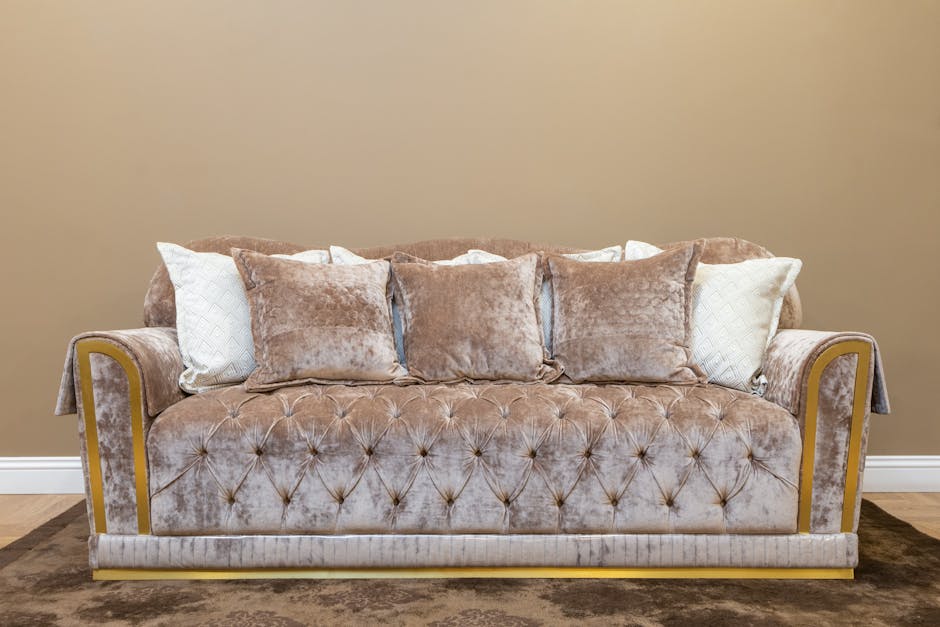 A plush, beige velvet sofa with deep button tufting in a Mayfair living room, decorated with multiple throw pillows in coordinating shades of cream and taupe, positioned against a plain, light tan wall. The sofa features rolled arms with wooden trim accents, and the seating area appears clean and well-maintained. A dark brown rug is visible beneath the sofa, and the room is evenly lit, highlighting the luxurious fabric and smooth surface of the furniture, in a setting that reflects high-quality upholstery cleaning and surface maintenance by Carpet Cleaners Mayfair.