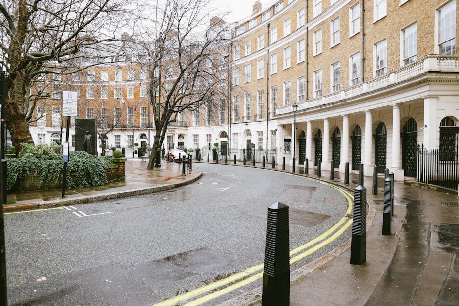 A city street in Mayfair W1K with row houses constructed from red brick, featuring multiple windows with white frames and decorative gables. The street has a section cordoned off with orange and white