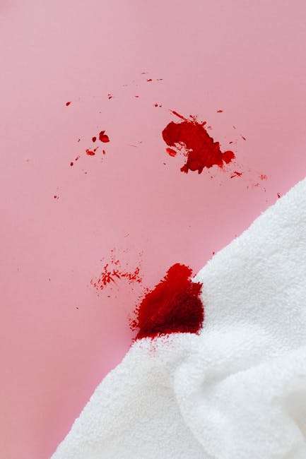 A close-up image of a pink surface, likely a wall or tabletop, showing a fresh red stain resembling spilled juice or dye, with scattered smaller splashes nearby. In the lower right corner, a white cloth partially covers the surface, suggesting a cleaning or blotting action. The lighting is neutral, highlighting the contrast between the bright pink background, the vivid red stain, and the white fabric. This image illustrates the need for surface cleaning and stain removal, as part of comprehensive domestic or commercial sanitisation and deep cleaning services offered by Carpet Cleaners Mayfair, demonstrating their expertise in same-day stain removal in Mayfair.
