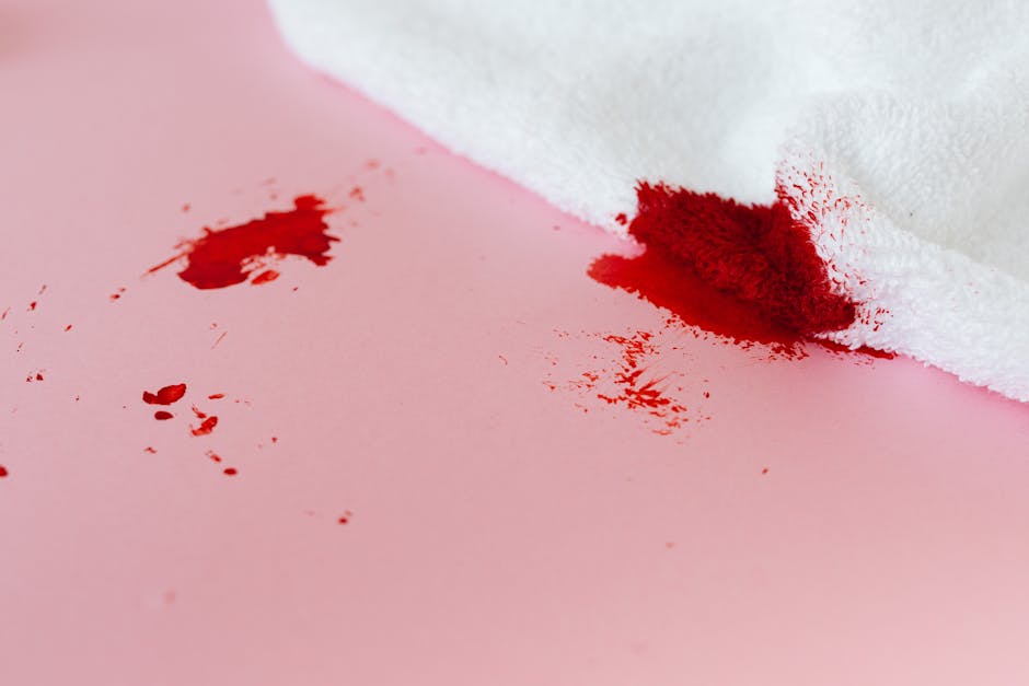 A close-up image of a pink surface, likely a wall or tabletop, showing a fresh red stain resembling spilled juice or dye, with scattered smaller splashes nearby. In the lower right corner, a white cloth partially covers the surface, suggesting a cleaning or blotting action. The lighting is neutral, highlighting the contrast between the bright pink background, the vivid red stain, and the white fabric. This image illustrates the need for surface cleaning and stain removal, as part of comprehensive domestic or commercial sanitisation and deep cleaning services offered by Carpet Cleaners Mayfair, demonstrating their expertise in same-day stain removal in Mayfair.