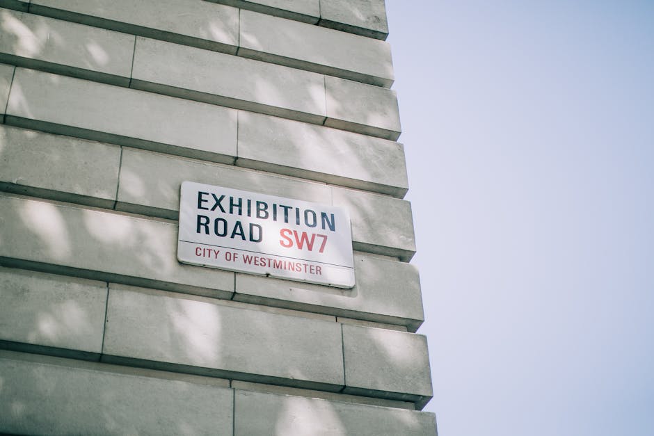 A close-up view of a building corner with a white street sign reading 'Exhibition Road SW7, City of Westminster' attached to a light-colored stone wall. The wall features horizontally aligned stone blocks with smooth surfaces, some faint shadows cast by nearby trees. The background includes a clear blue sky, and the lighting appears natural during daytime. The image highlights the clean and well-maintained façade, emphasizing the urban context, as part of content related to surface cleaning and maintenance by Carpet Cleaners Mayfair, aligning with Westminster Council regulations for waste management from Mayfair cleans.