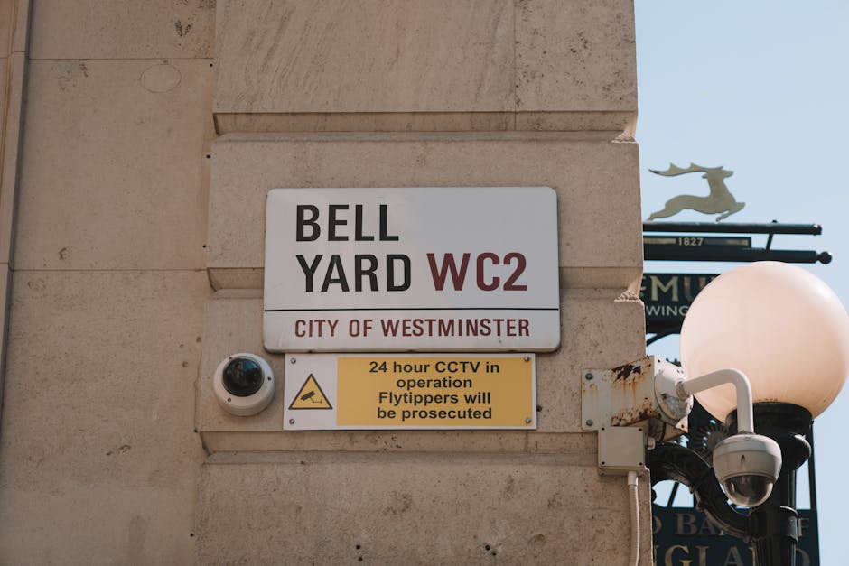 A close-up photograph of a beige brick wall featuring a metal street sign that reads 'BELL YARD WC2 CITY OF WESTMINSTER.' Below the sign, there is a yellow warning notice indicating '24 hour CCTV in operation Flytippers will be prosecuted,' with a black surveillance camera mounted nearby. To the right, a black lamppost with a white, spherical lamp is visible, set against a background of a clear blue sky. The image captures the exterior of an urban building, emphasizing security features and signage related to waste regulations and surveillance in Westminster.