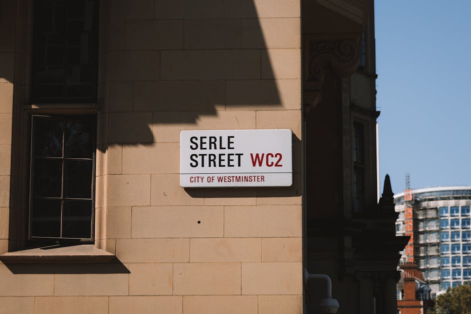 A close-up view of a building corner with a white street sign reading 'Exhibition Road SW7, City of Westminster' attached to a light-colored stone wall. The wall features horizontally aligned stone blocks with smooth surfaces, some faint shadows cast by nearby trees. The background includes a clear blue sky, and the lighting appears natural during daytime. The image highlights the clean and well-maintained façade, emphasizing the urban context, as part of content related to surface cleaning and maintenance by Carpet Cleaners Mayfair, aligning with Westminster Council regulations for waste management from Mayfair cleans.