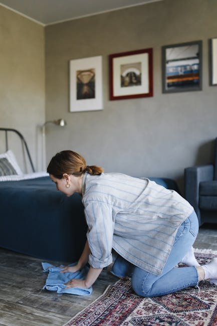 A woman with brown hair tied back, wearing a light blue and white striped shirt and blue jeans, is kneeling on a patterned area rug while cleaning a wooden floor with a blue cloth. The room features a bed with a white pillow and dark bedding against a grey wall decorated with three framed artworks. A grey armchair is visible in the background, and natural light illuminates the space, highlighting the cleanliness and dust-free condition of the room. This scene illustrates domestic cleaning practices, emphasizing surface cleaning and maintaining hygiene, as part of professional services offered by Carpet Cleaners Mayfair.
