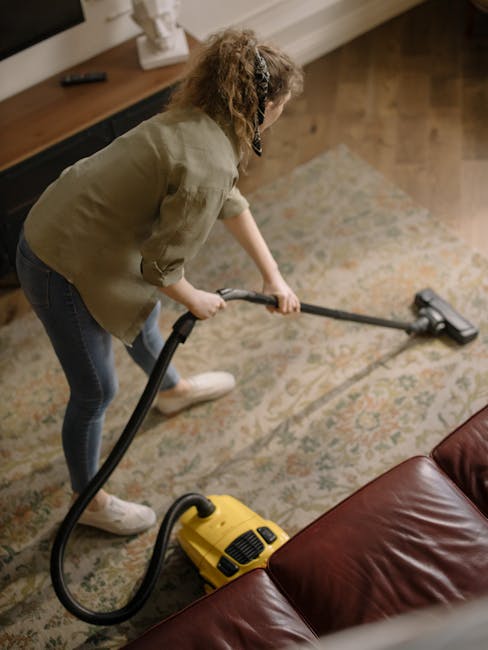 A woman with curly blond hair, wearing a beige jacket, blue jeans, and white shoes, is engaged in surface cleaning with a vacuum cleaner on a patterned, beige area rug in a living room. The vacuum cleaner is yellow with a black hose and nozzle, which she is directing over the rug's surface. She is standing beside a brown leather sofa, and in the background, there is a wooden console table with a white decorative object and other small items. The room is well-lit with natural light, highlighting the clean and tidy appearance of the space, which also features wooden flooring adjacent to the rug. This scene exemplifies deep cleaning and sanitisation practices typical of domestic cleaning services provided by Carpet Cleaners Mayfair, with an emphasis on surface hygiene and maintaining a fresh, hygienic home environment.